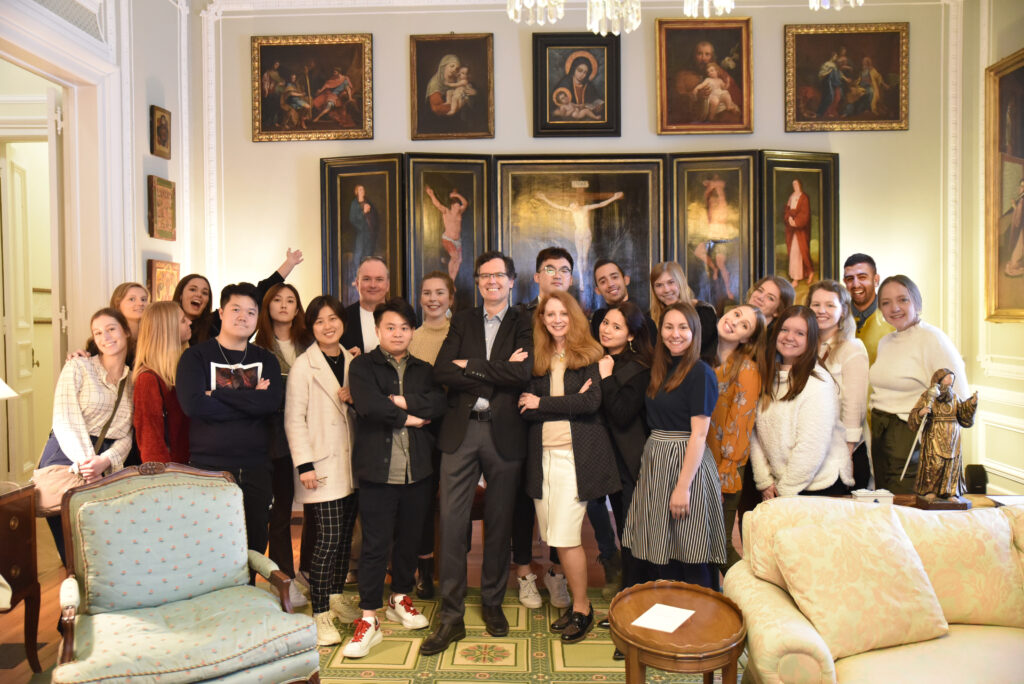 Color photograph, group of men and women standing, and smiling, looking toward the viewer; they are standing in a living room with floor carpet, chandelier, and in front of a wall with several paintings.