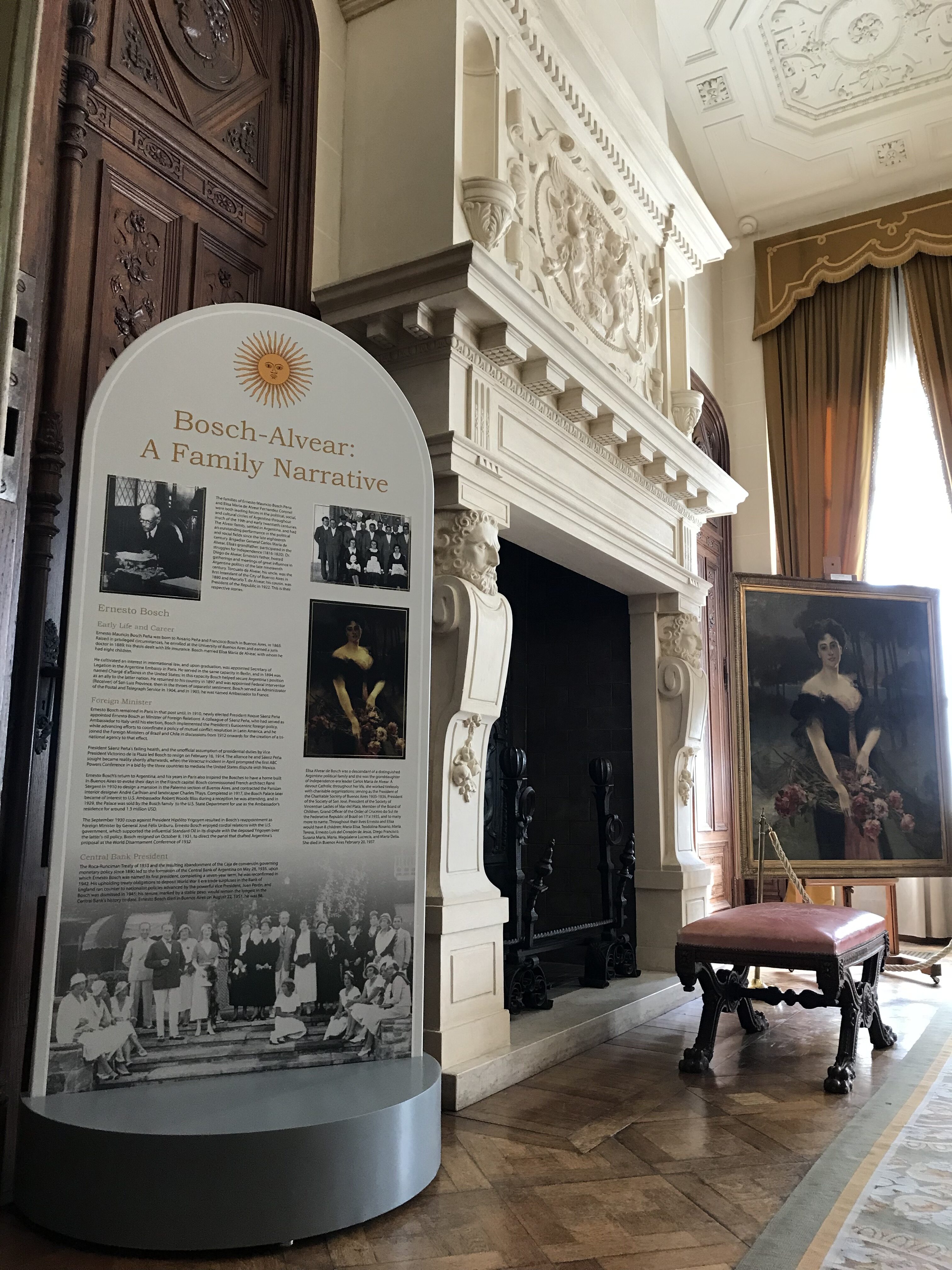 Color photograph, museum display panel and portrait placed to either side of fireplace with decoratively carved surround and overmantel.