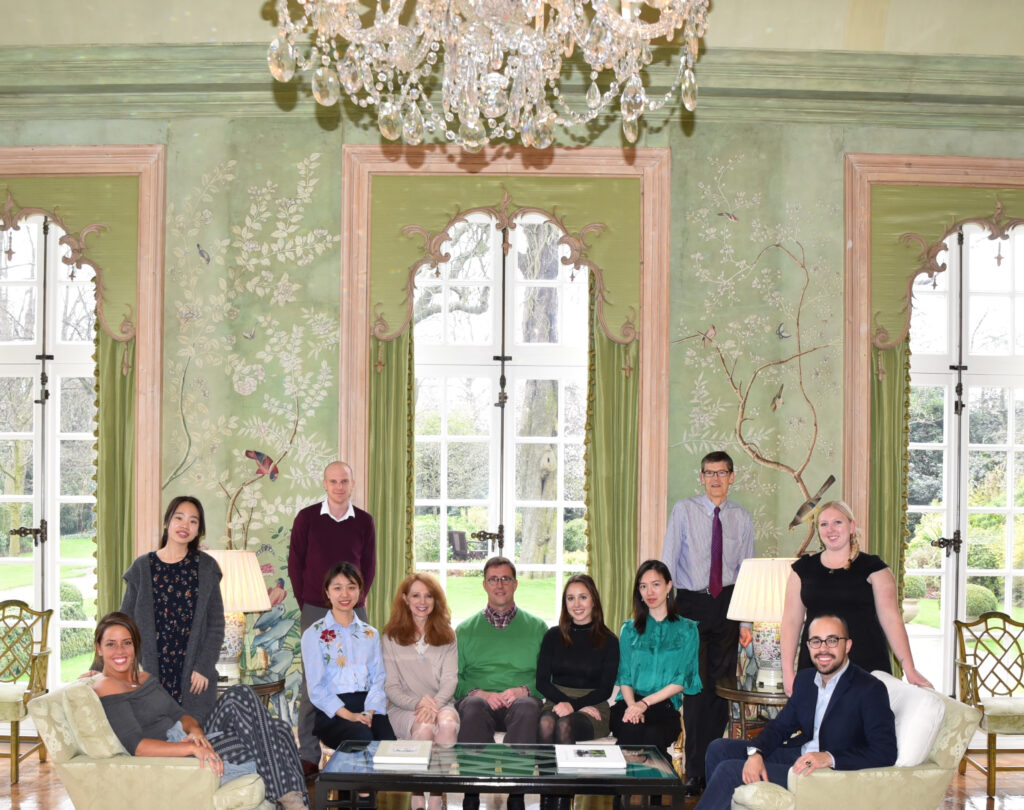 Color photograph, men and women sitting and standing in the Garden Room of Winfield House, London; the wallpaper is hand-painted and has a green background.