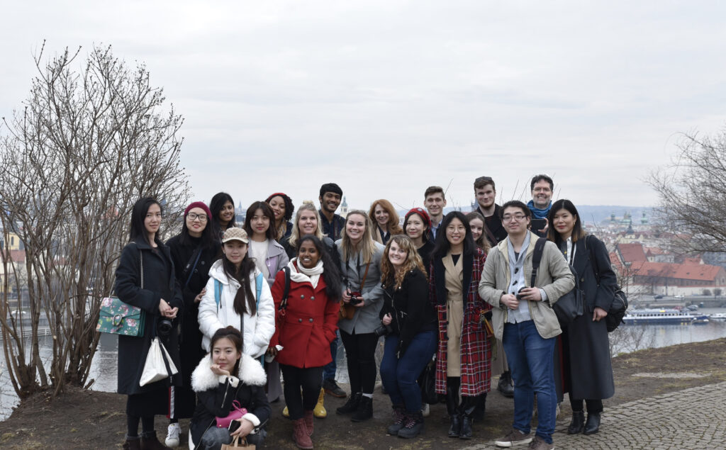 Color photograph, group of men and women posing for a picture. They are outside and standing near a river embankment - the brick paved path is in front of the them; the water is below and red roof tops and church spires of the city beyond.