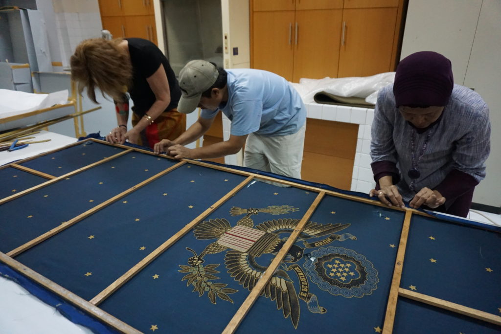 Color photograph, three people leaning over a framed tapestry; The fabric is blue with an eagle and small gold stars.