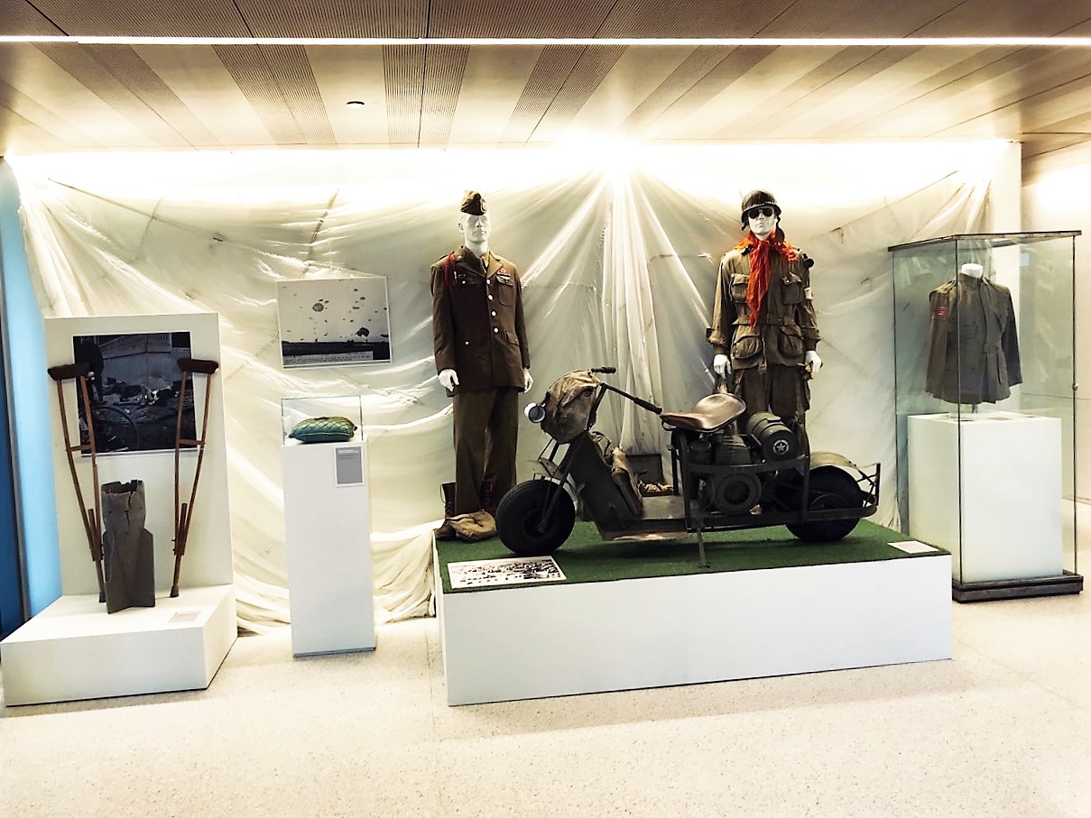 Color photograph, exhibit diplay platform and cases for a WWII era motorbike, uniforms, and crutches with a white parachute backdrop