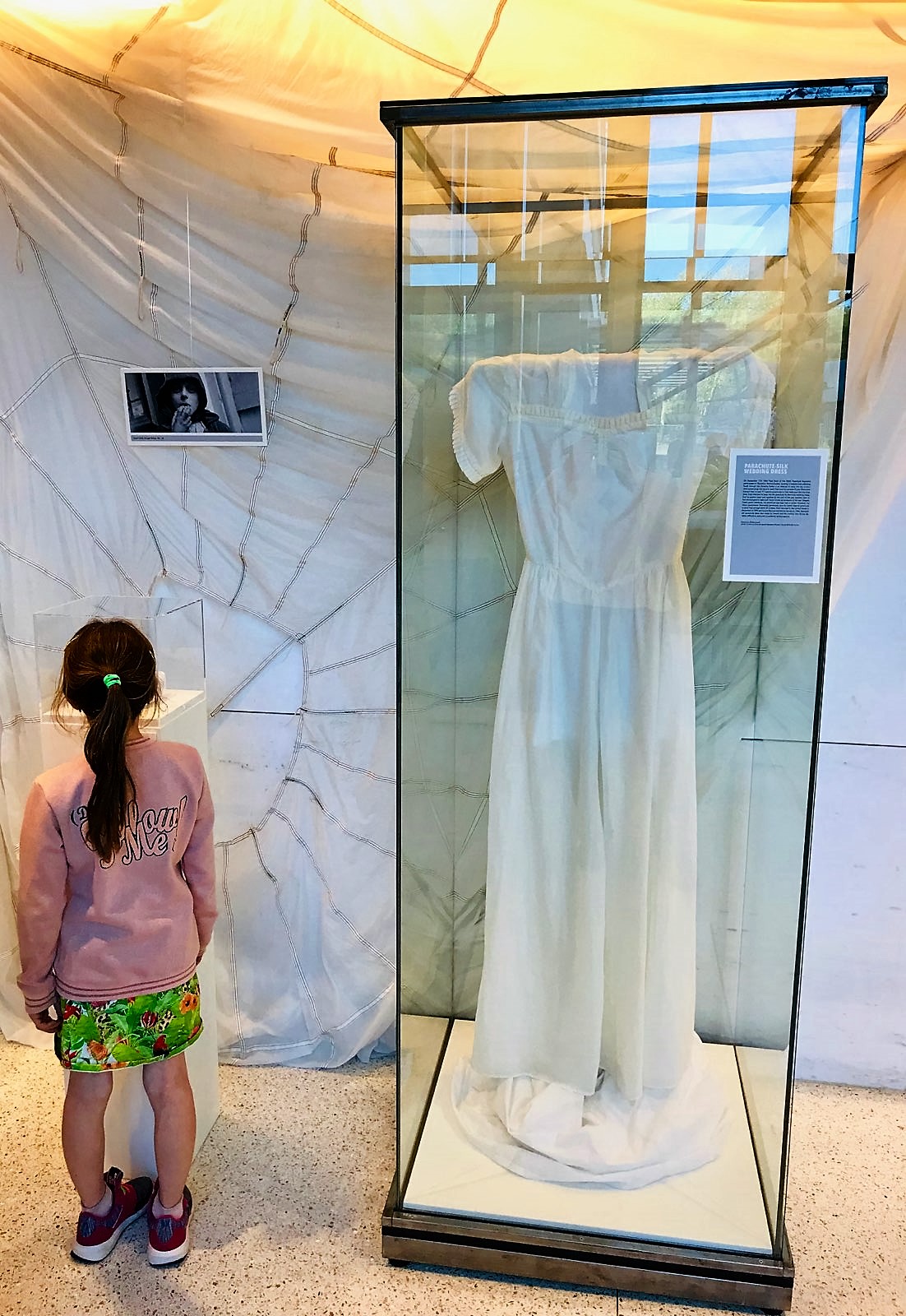 Color photograph, young girl looking at museum diplay text and white dress. A white parachute creates the backdrop.