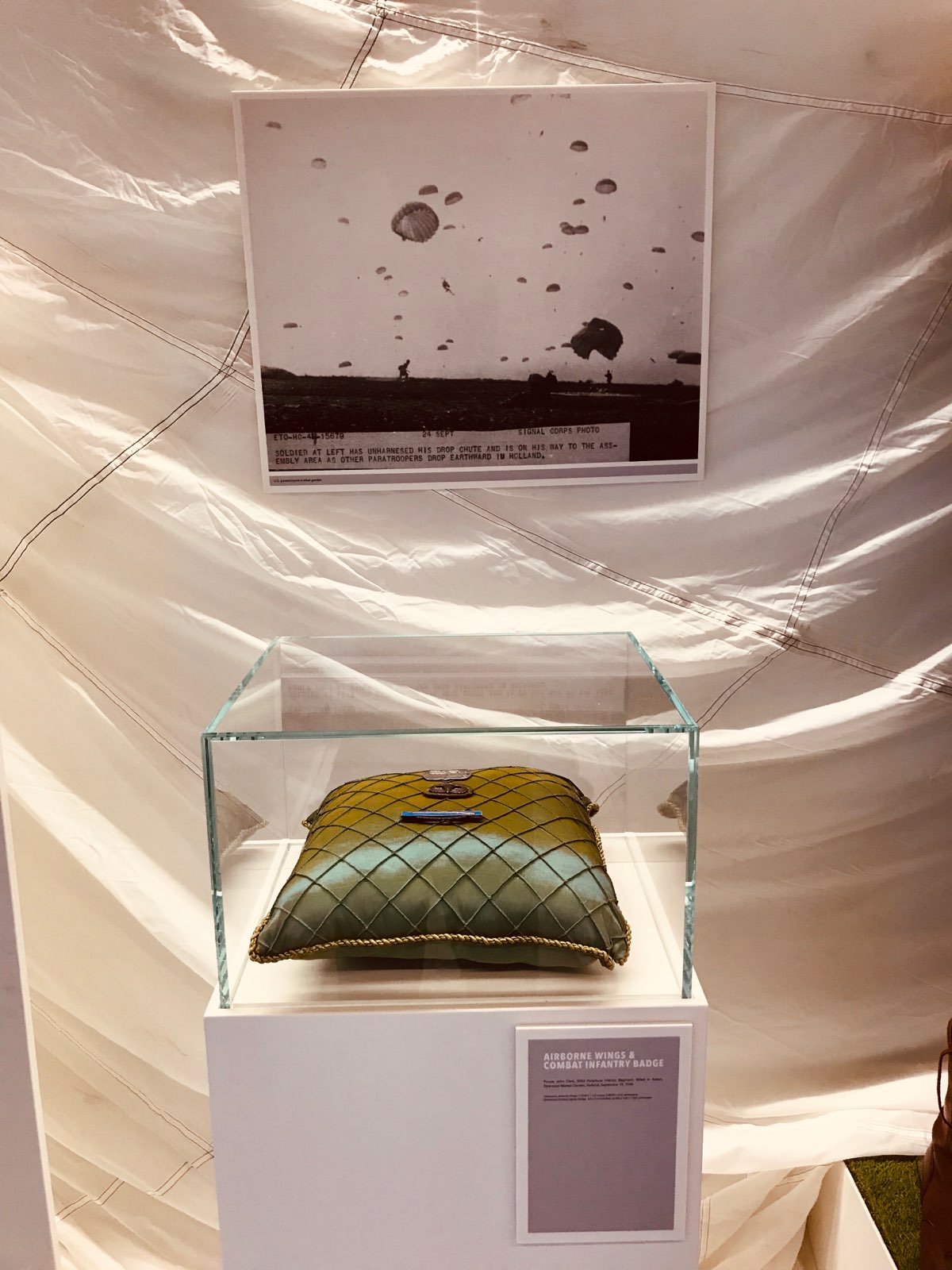 Color photograph, museum display of airbourne wings and combat infantry badges on a green pillow. A black and white photograph is in the background.