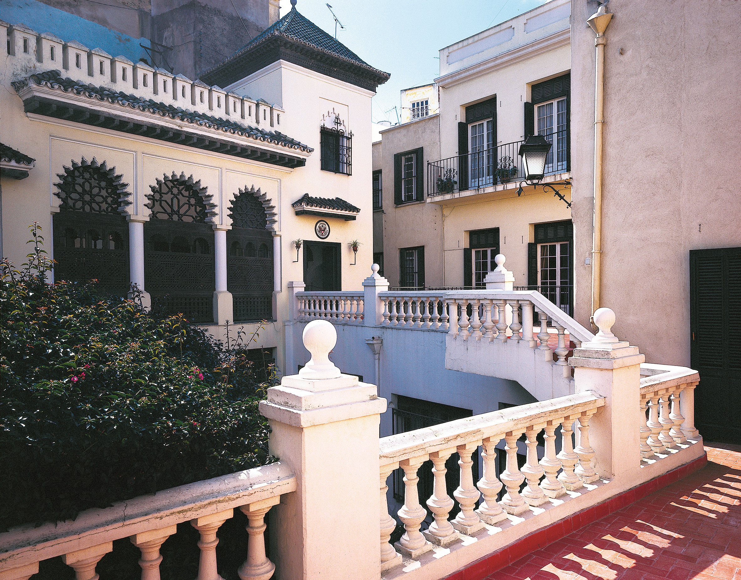 Color photograph, Color photograph, view from balcony with red tile and white balustrade with bulbous balusters across an open courtyard to a loggia. The entrance to the loggia to the side and a small, tiled hood covers the door. The seal of the United States is over the door.