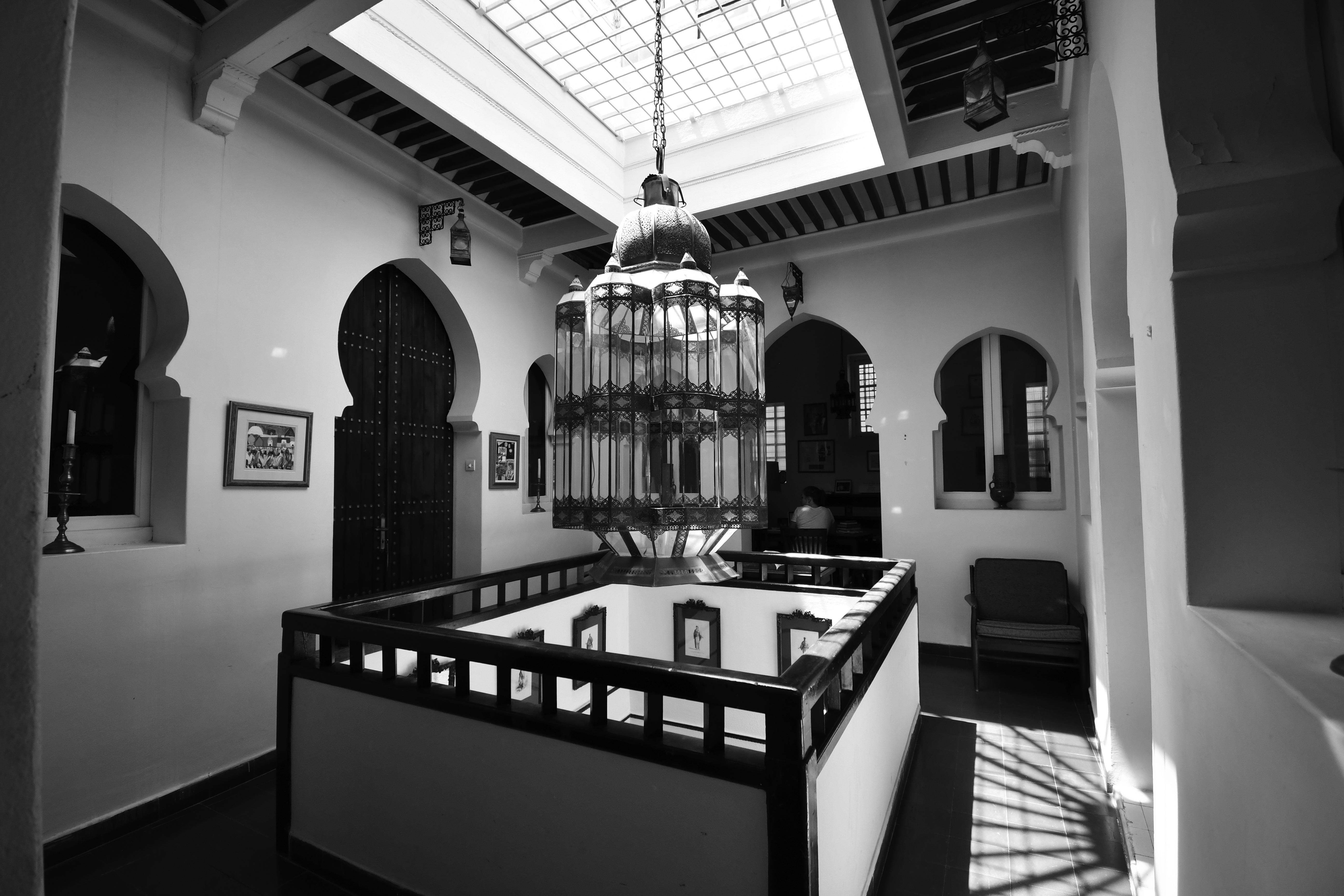 Black and white photograph, view across interior space lit by a skylight with open well to light the floors below; the open area is famed by low walls with a wood railing. The doors to spaces opening off this central area have Moorish arches. A chandelier is in the foreground.