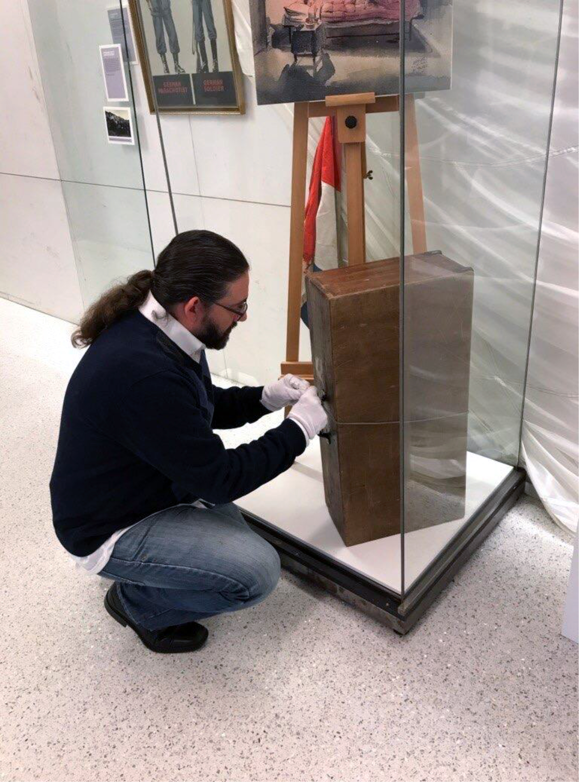 Color photograph, man assembling museum display of wooden suitcase and images on an easel.