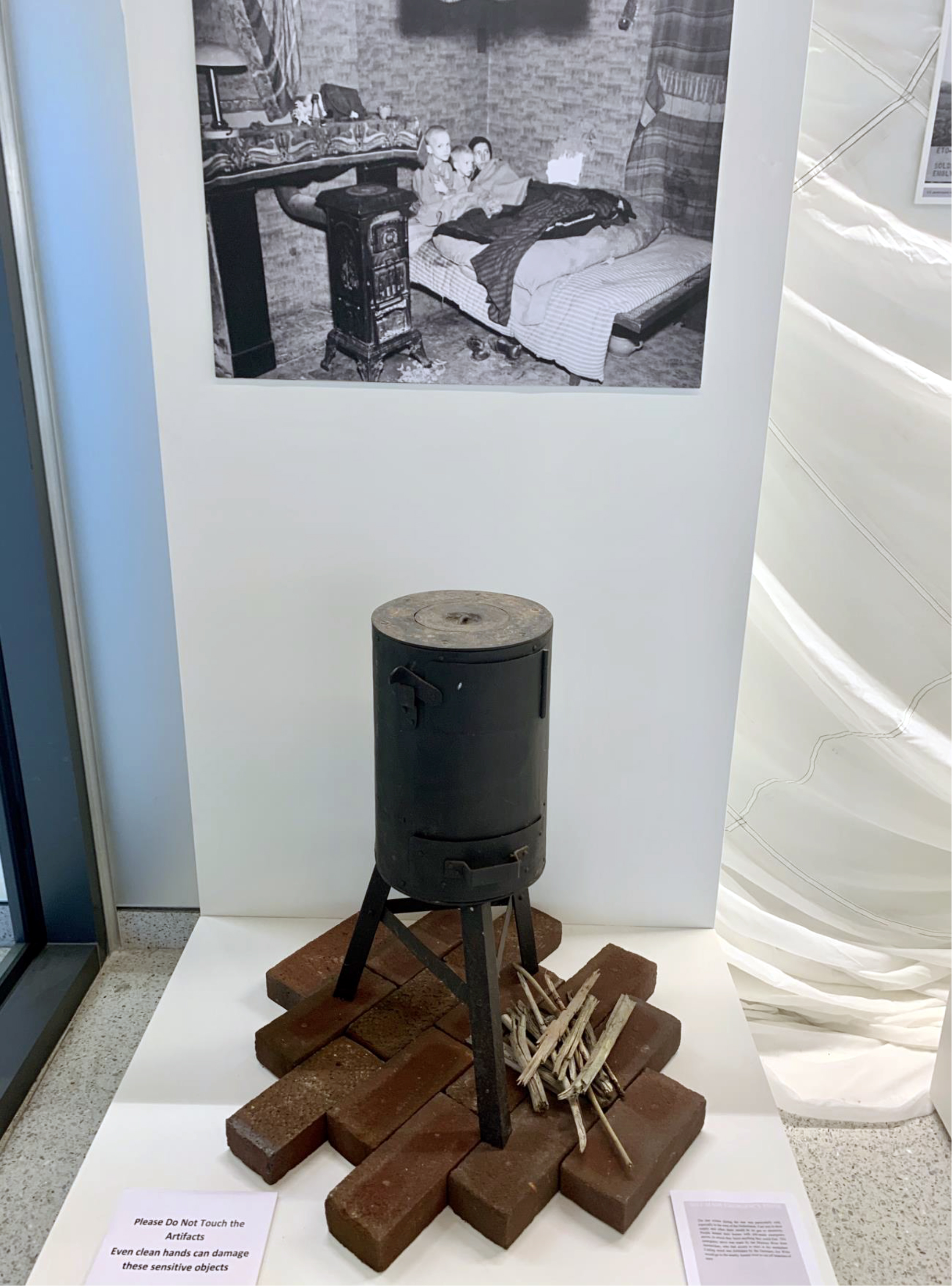 Color photograph, museum display of camp stove on loose bricks, with black and white photograph in background