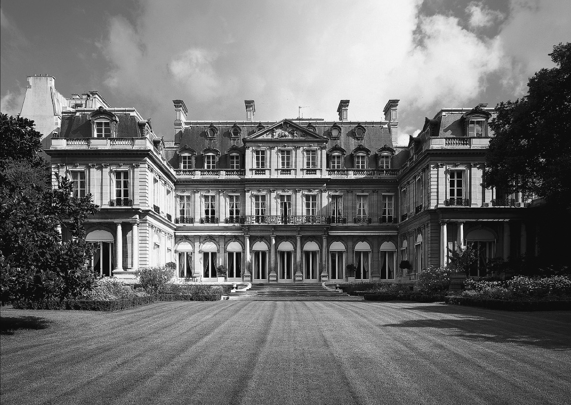 Black and white photograph, view across green grass to a U-shaped building with classical details and symmetrically placed window and door openings.