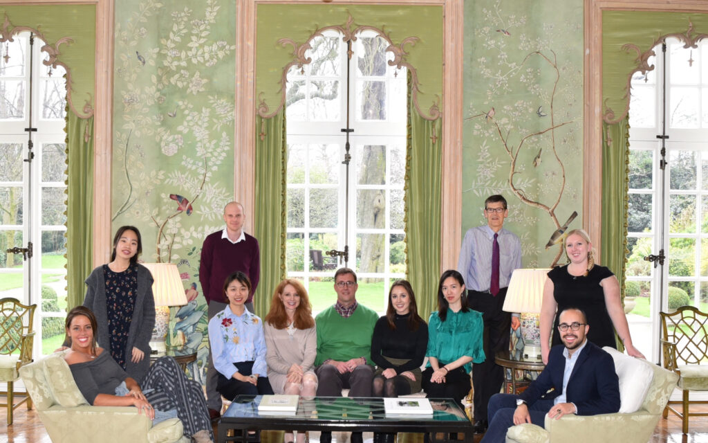 Color photograph, men and women sitting and standing in the Garden Room of Winfield House, London; the wallpaper is hand-painted and has a green background.