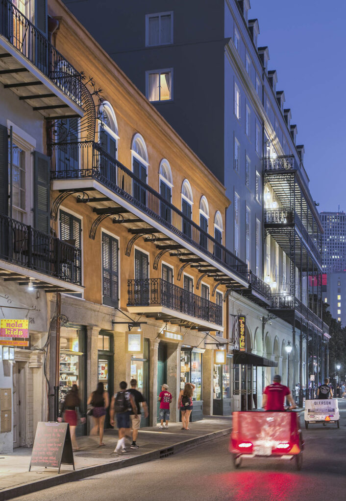 Color photograph, New Orleans streetscape at night and perspective view of a three-story building with ground-level retail and an iron balcony at the third floor.