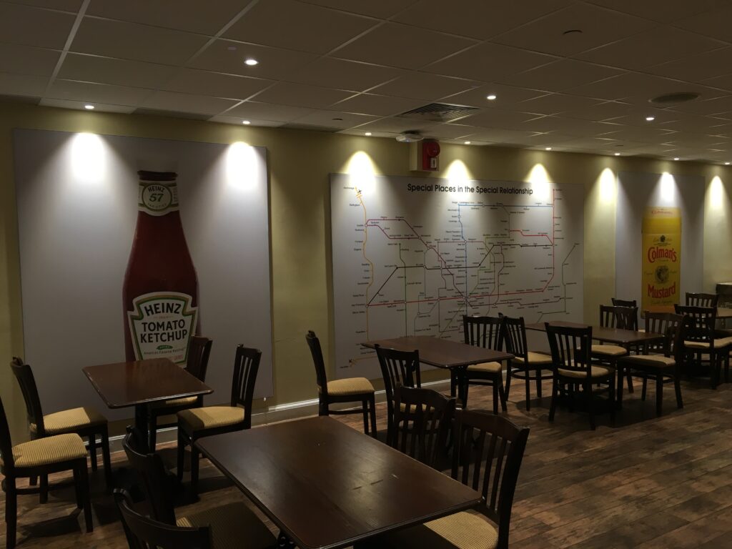 Color photograph, tables and chairs in a room with large map and a poster image of bottle of ketchup on the wall in background