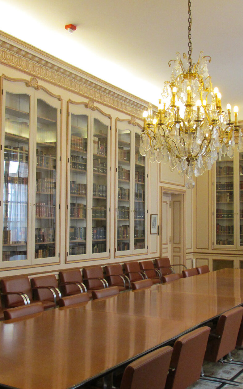 Color photograph, interior of a room looking diagonally across a long conference table and chairs to a recessed doorway. There is a chandelier above the table. The walls are painted and have built-in bookcases with glass fronts.